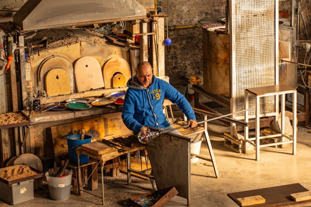 Glassblower crafting art in traditional Venetian workshop with furnaces and tools.